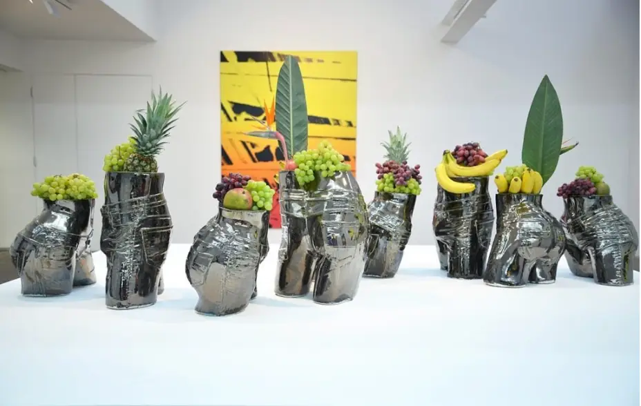 A sculptural installation featuring eight metalic figures from the waist to the upper legs. Each figure is topped with a combination of grapes, bananas, mango, and pineapples.